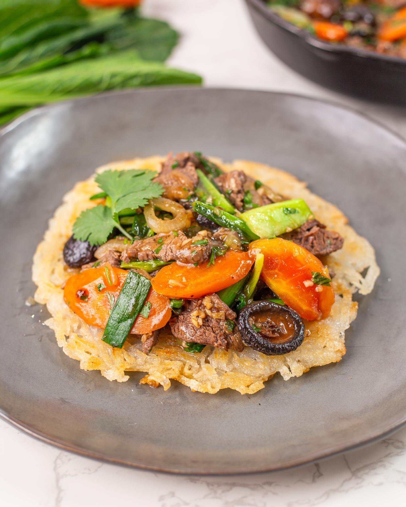 Gray plate with crispy fried rice noodles that form a round flat disk, topped with stir-fried beef and different types of vegetables.